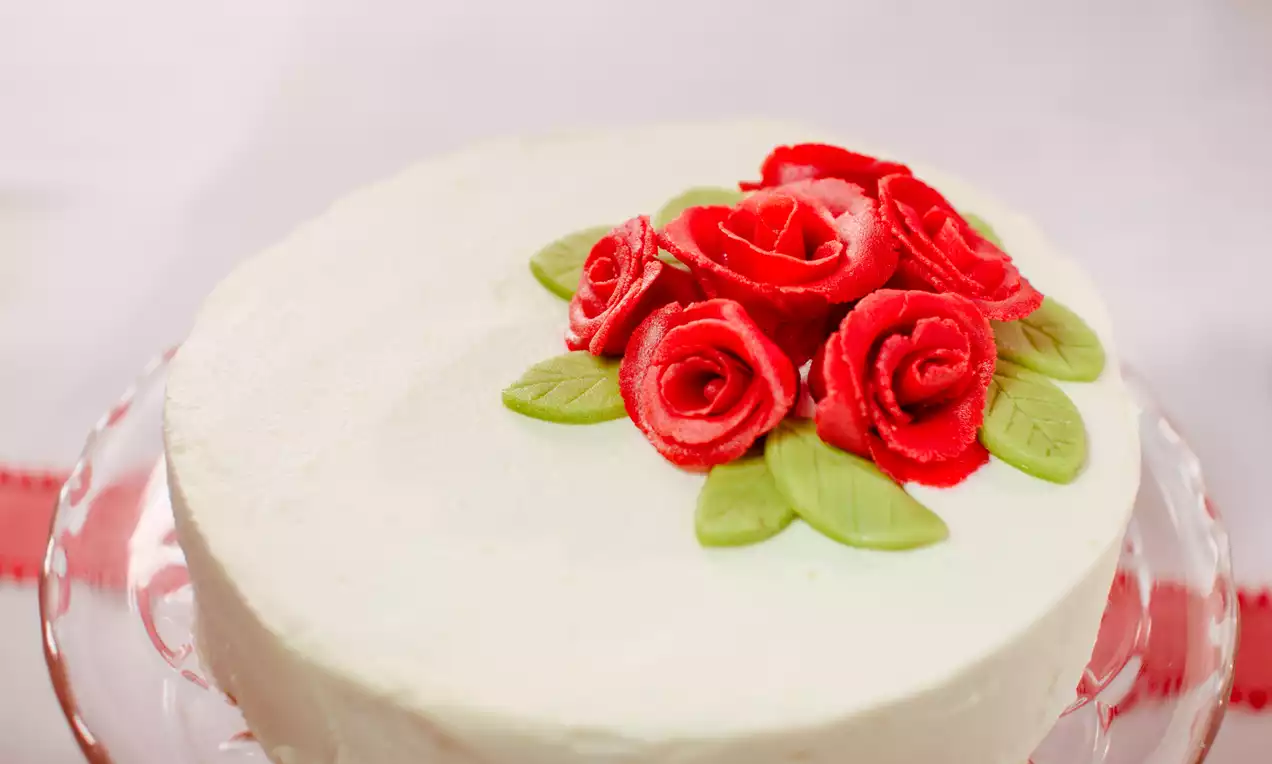 Weiße Topfen-Zitronen-Obers-Torte mit glatter Oberfläche, dekoriert mit roten Zuckerrosen und grünen Blättern auf einer Glasplatte