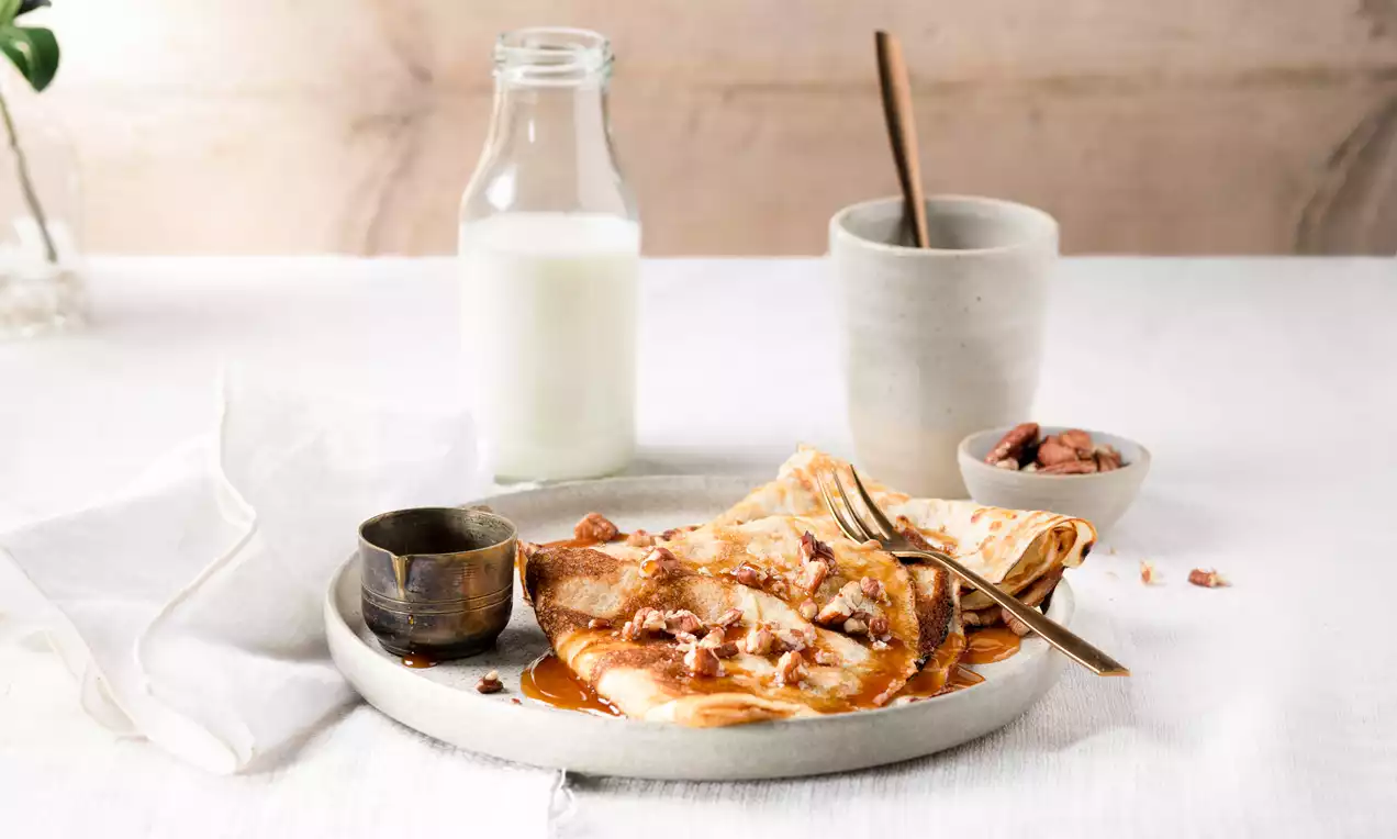 Crêpes dorées légèrement pliées, nappées de caramel ambré, parsemées de noix croquantes, servies sur une assiette beige avec un pot de caramel et un verre de lait