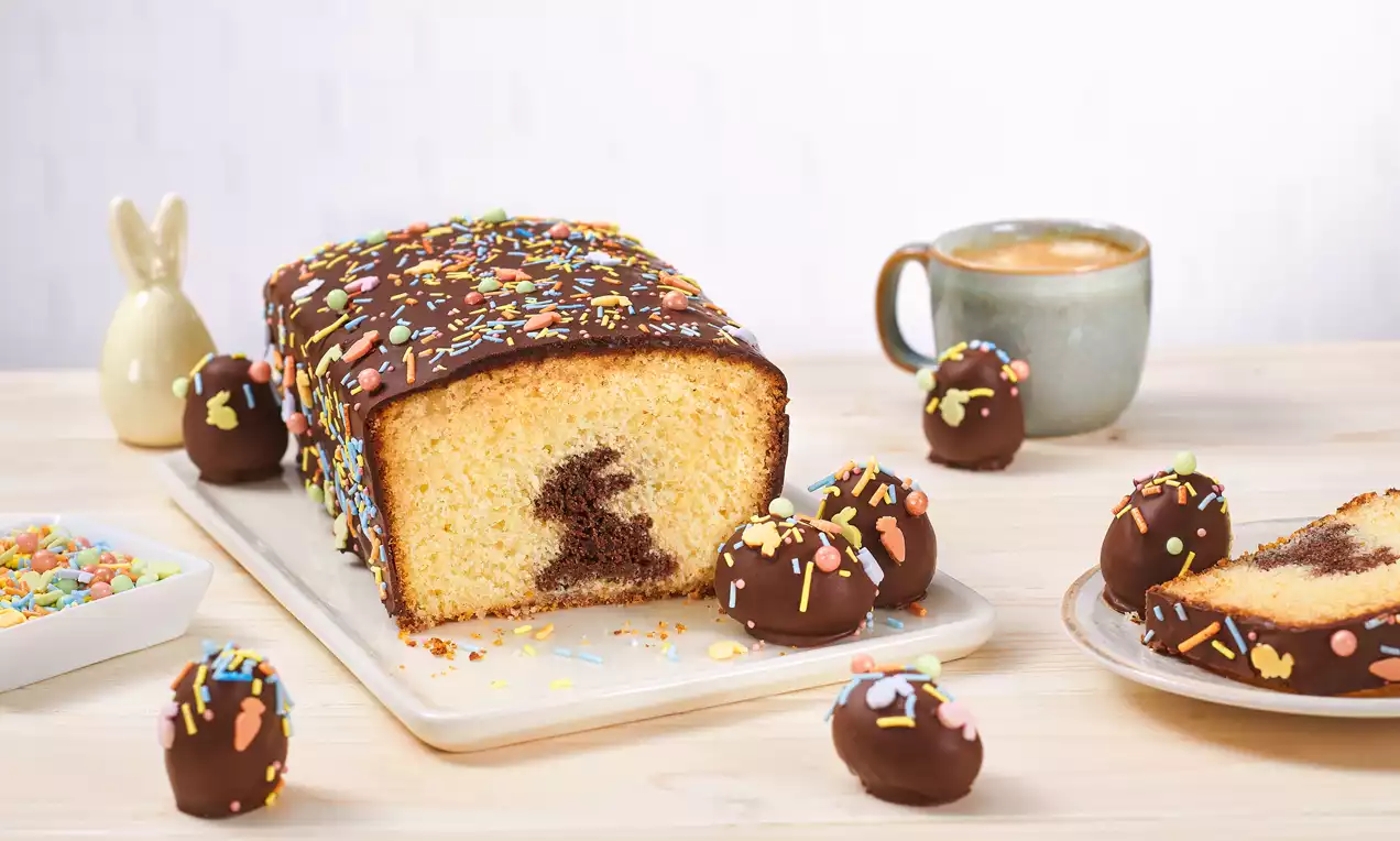 Oster-Kastenkuchen mit Schokoglasur und bunten Streuseln, innen Häschenmotiv, dekoriert mit Schokopralinen auf hellem Holztisch