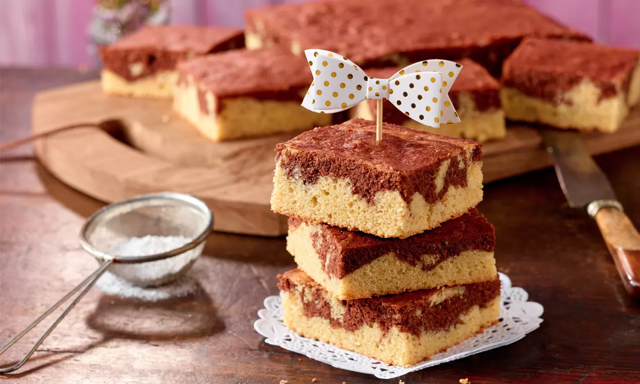 Drei Stücke Schoko-Bananen-Marmorkuchen in Quadratform mit goldgelbem Boden und dunkler Marmorierung, gestapelt auf einer weißen Spitzendecke