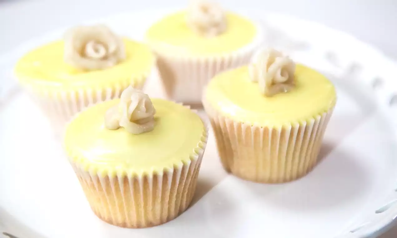 Four pale yellow cupcakes with glossy lemon icing, topped with white chocolate rose decorations, arranged on a white plate