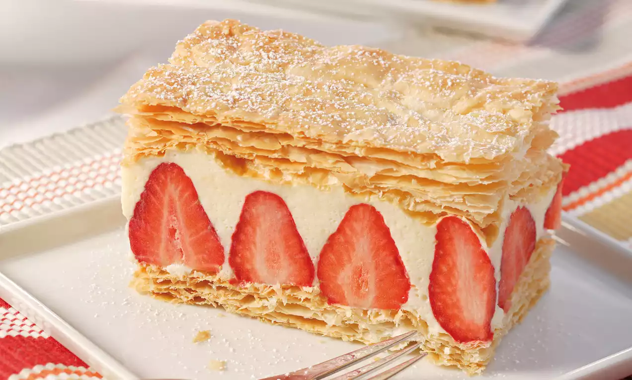 Goldbraune Blätterteigschichten mit cremiger Füllung und eingebetteten roten Erdbeerscheiben, bestäubt mit Puderzucker auf weißem Teller