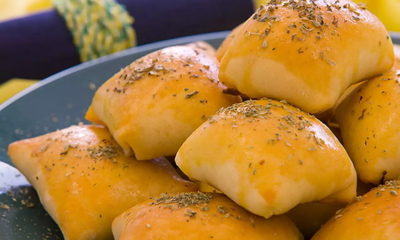 Mini pães dourados em formato quadrado com superfície brilhante e salpicada de ervas sobre prato azul com guardanapo decorativo ao fundo