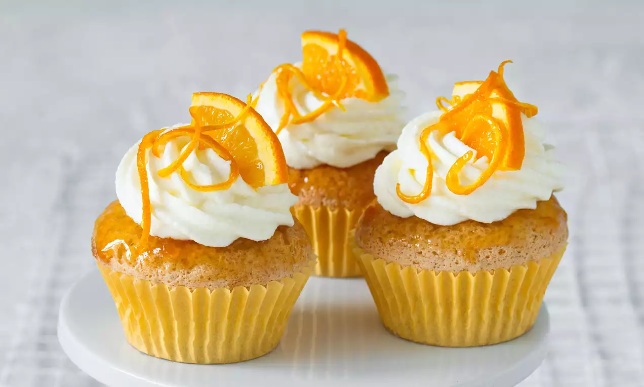 Pomerančové cupcakes v žltých košíčkoch s nadýchanou bielou šľahačkou ozdobenou špirálami pomarančovej kôry na bielom podnose