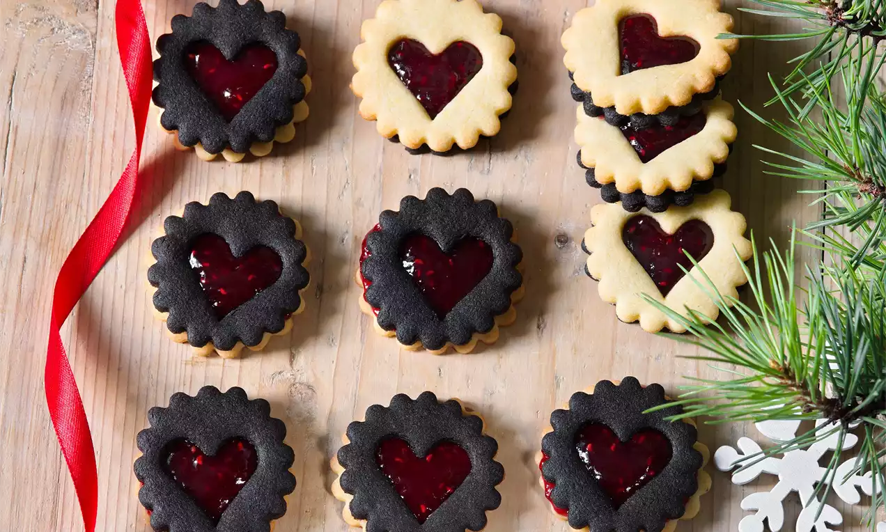 Linecká kolečka srdcového tvaru s hladkým malinovým džemem, střídající světlé a tmavé korpusy, na dřevěném stole s vánoční výzdobou