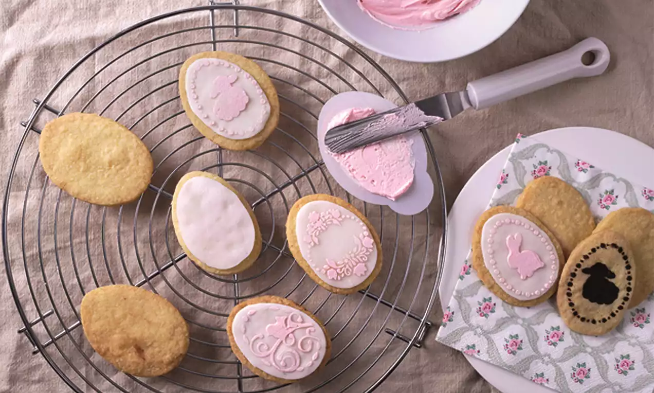 Goldgelbe, ovale Kekse mit zartrosa Dekor und filigranen Mustern, teils auf einem Kuchengitter, teils auf einer Serviette arrangiert