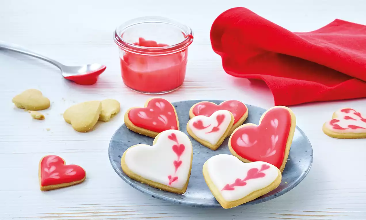 Herzförmige Kekse mit roter und weißer Zuckerglasur auf grauem Teller, dekoriert mit Herzmustern, daneben rotes Tuch und Glasur im Glas