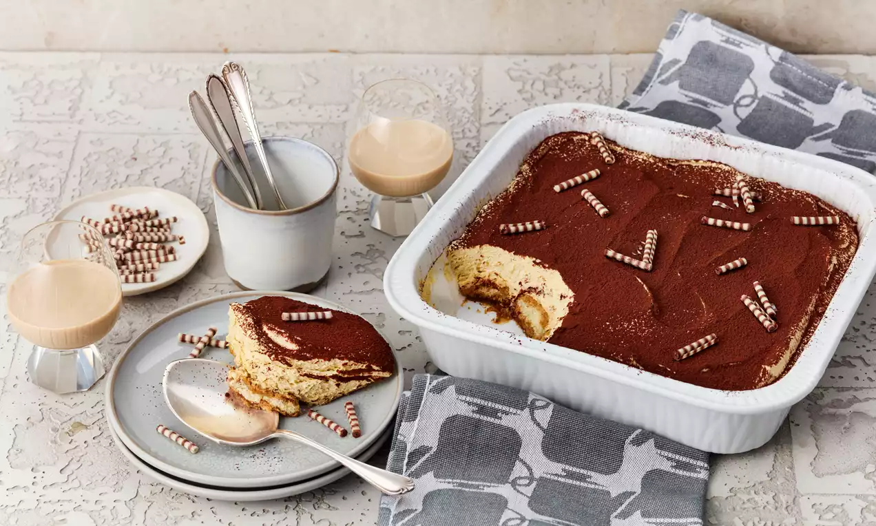 Baileys-Tiramisu mit hellgelber Creme, Kakaopulver und eingerollter Deko in weißer Form, daneben ein Teller mit serviertem Stück
