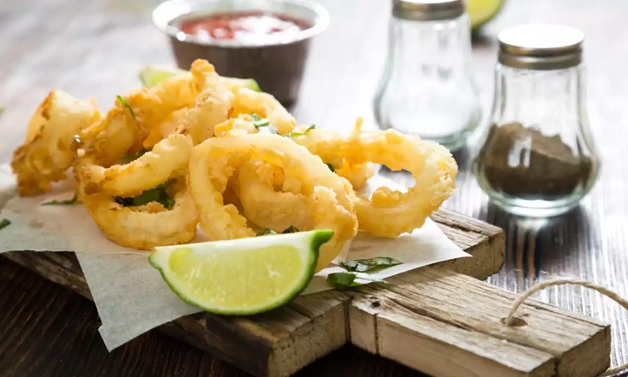 Golden, crispy calamari rings arranged on parchment on a rustic wooden board, garnished with fresh herbs and a lime wedge