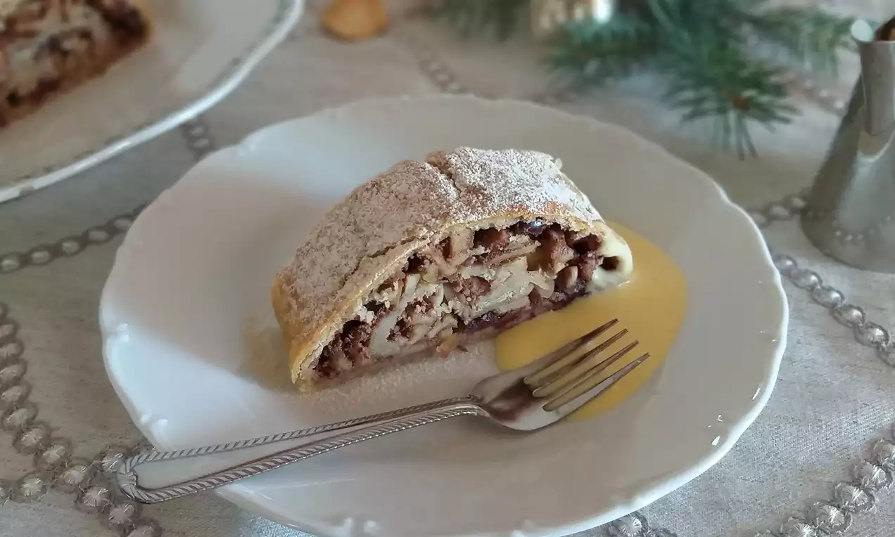 Plátok zlatohnedého jablkového štrúdľa s práškovým cukrom, položený na bielom tanieri s krémovou vanilkovou omáčkou a vidličkou, na sviatočnom obruse.