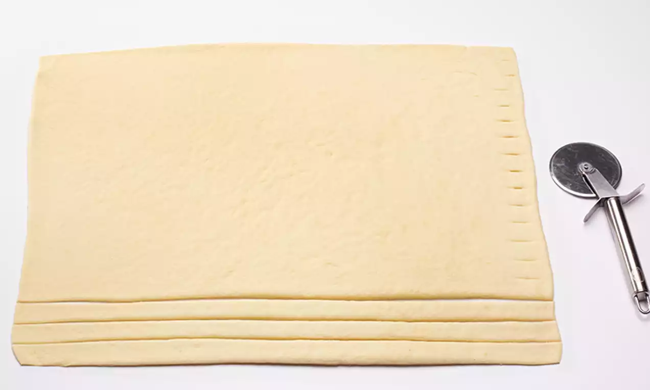 Pâte feuilletée rectangulaire d'un jaune pâle avec une texture lisse et légèrement marquée, accompagnée d'une roulette métallique brillante