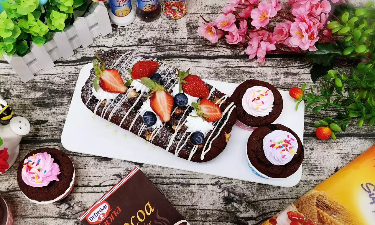 Super moist chocolate cake dark and glossy, topped with strawberries, blueberries, white drizzle and mint leaves on a white tray surrounded by pink flowers