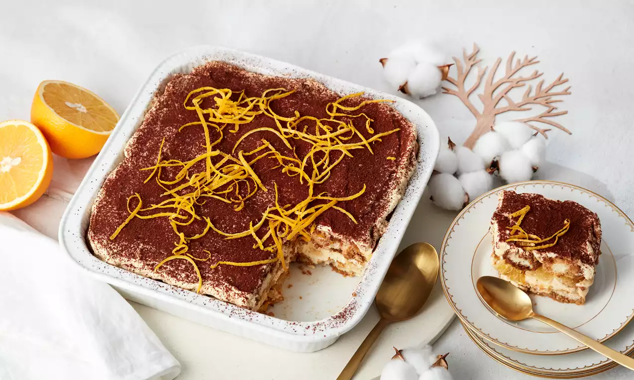 Orangen-Tiramisu mit braunem Kakaopulver und leuchtenden Orangenzesten in weißer Form, daneben ein Stück auf einem Teller mit goldenen Besteck