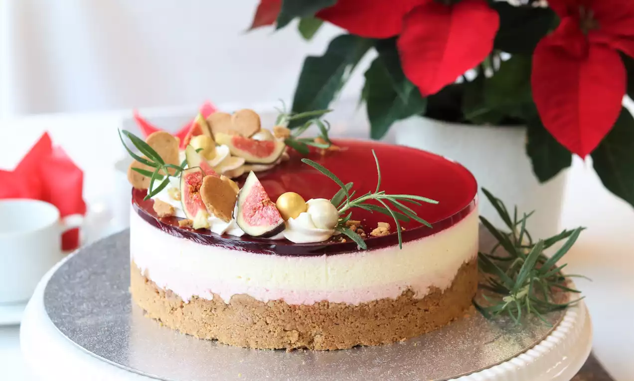 Juleostekake med brun kjeksbunn, lag av hvit og rosa ostekrem, rød gløgggele og pynt av fikener, rosmarin og gullkuler på et kakefat