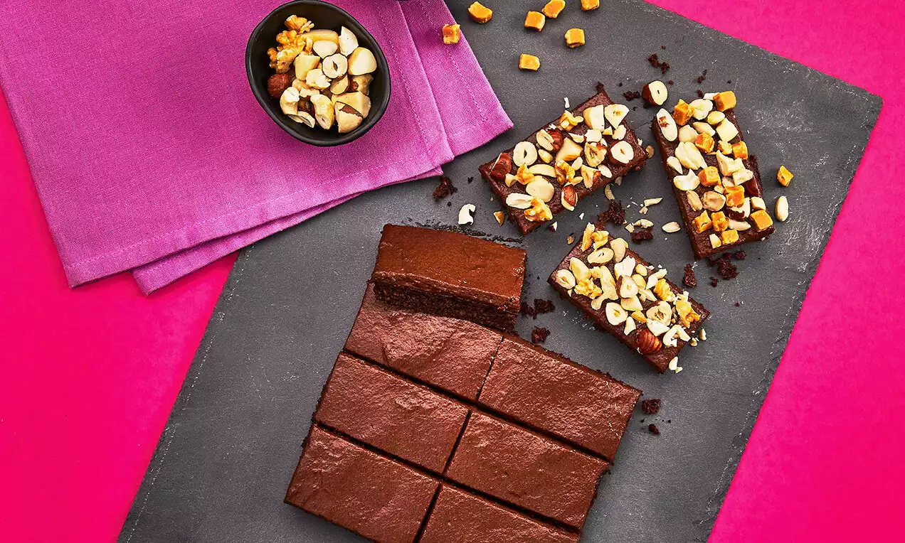 Tmavohnedé brownie s lesklým povrchom, rozkrájané na pravidelné štvorce, ozdobené kúskami orechov a sušeného ovocia na čiernej tácke