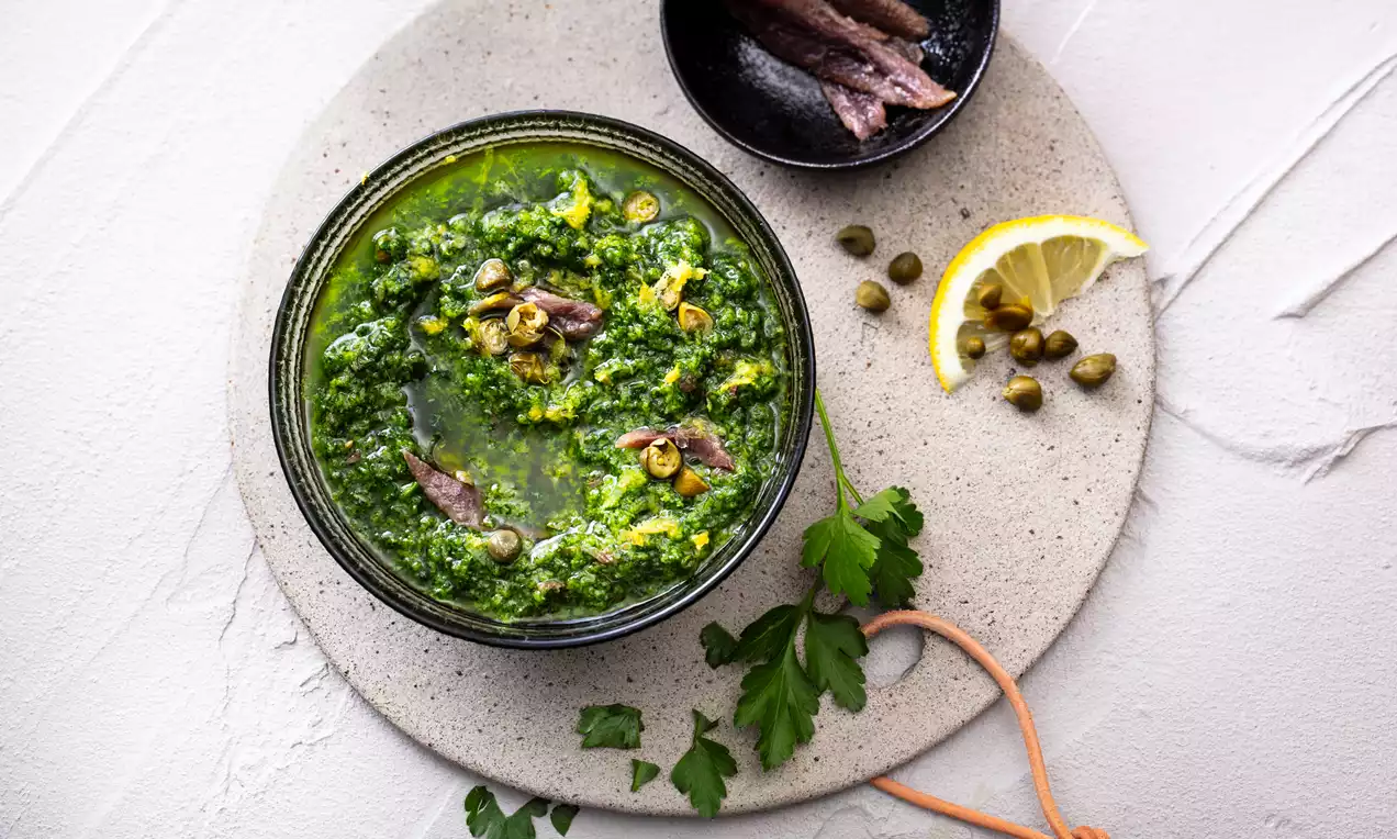 Groene salsa verde in een zwart schaaltje met stukjes ansjovis, kappertjes en citroenzeste, geserveerd op een lichtgrijze stenen plaat