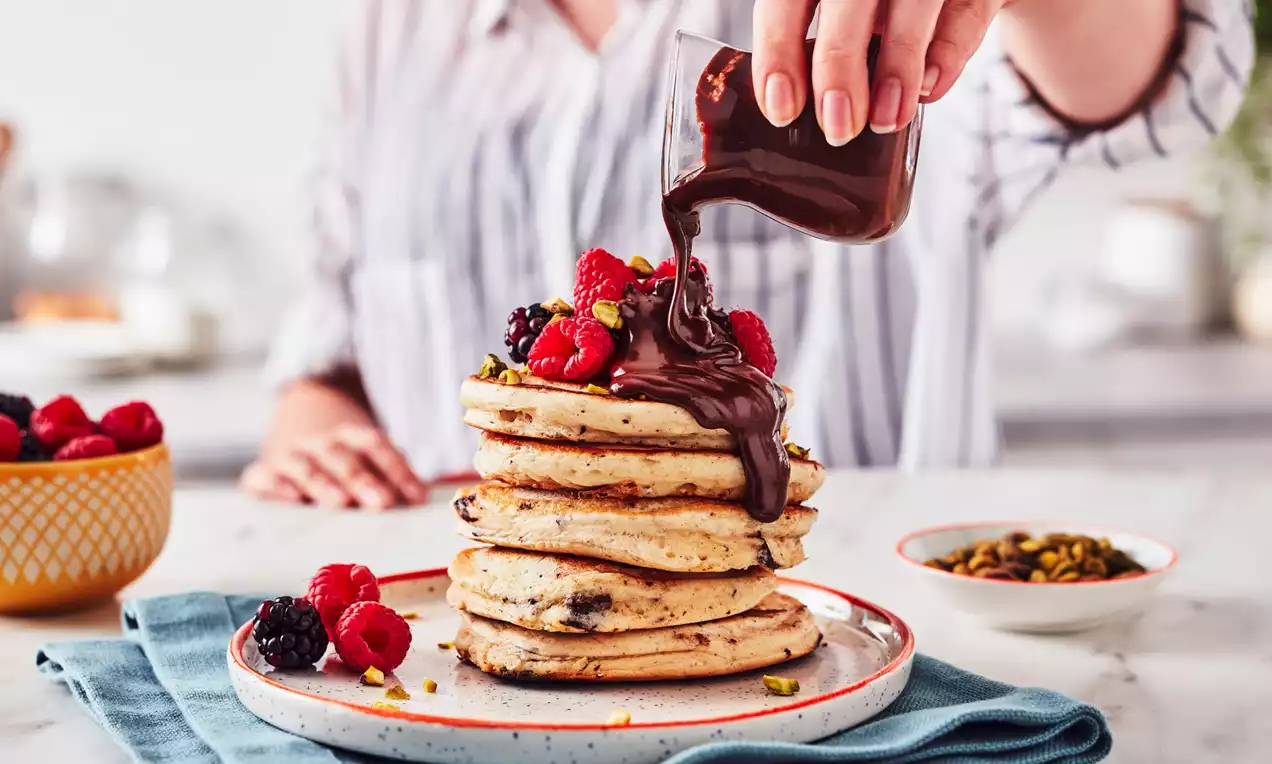 Üst üste dizilmiş altın rengi vegan pankekler, dökülen erimiş çikolata, taze frambuaz, böğürtlen ve antep fıstığı ile süslenmiş