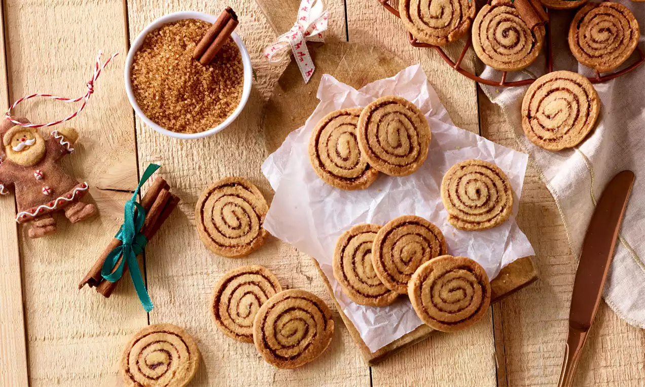 Goldbraune Zimtschnecken mit spiralförmiger Musterung auf weißem Backpapier, umgeben von braunem Zucker, Zimtstangen und Dekoration