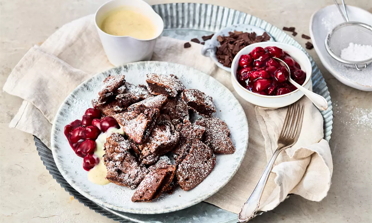 Schoko-Kaiserschmarrn in unregelmäßigen Stücken, dunkelbraun, mit Puderzucker bestäubt, serviert mit Vanillesoße und roten Kirschen auf einem Teller