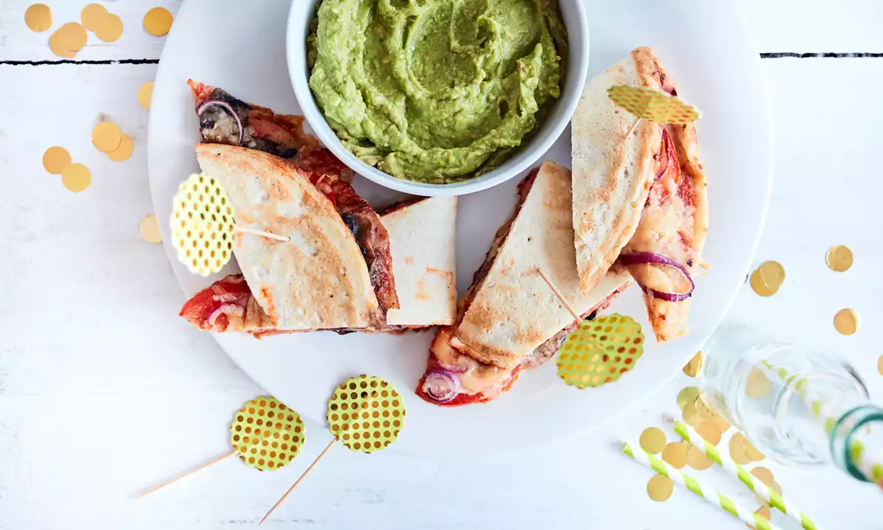 Driehoekige pizza-quesadillastukken met goudbruine korst en gesmolten kaas op een wit bord met een groen guacamoledip in het midden
