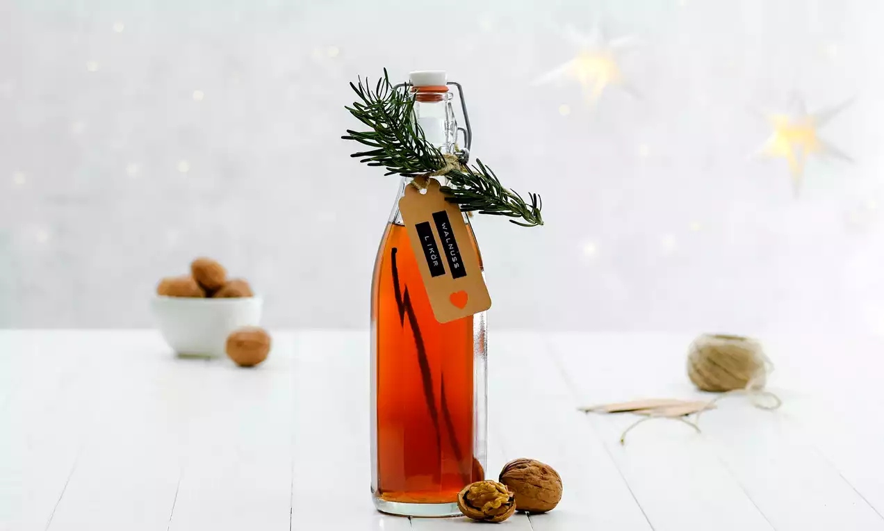 Staklena boca jantarnosmeđe tekućine s grančicom ružmarina i etiketom, okružena orasima na bijeloj drvenoj površini