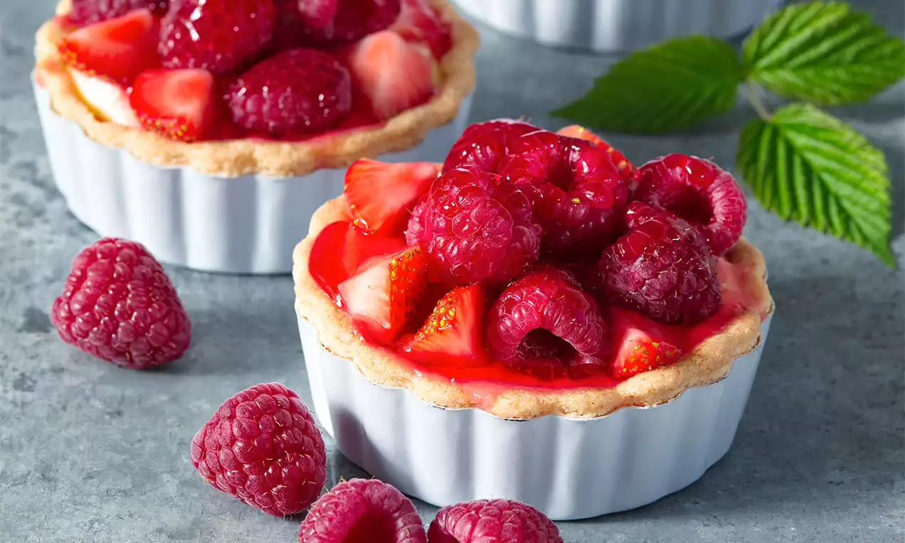 Bezlepkové košíčky v bielych keramických formičkách s jemne zlatistým okrajom, plnené krémom, malinami a jahodami na šedom povrchu