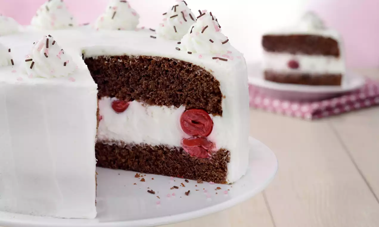 Czekoladowy tort Wuzetka z białym kremem jogurtowym, wiśniami w środku, udekorowany bitą śmietaną i czekoladową posypką na białym talerzu