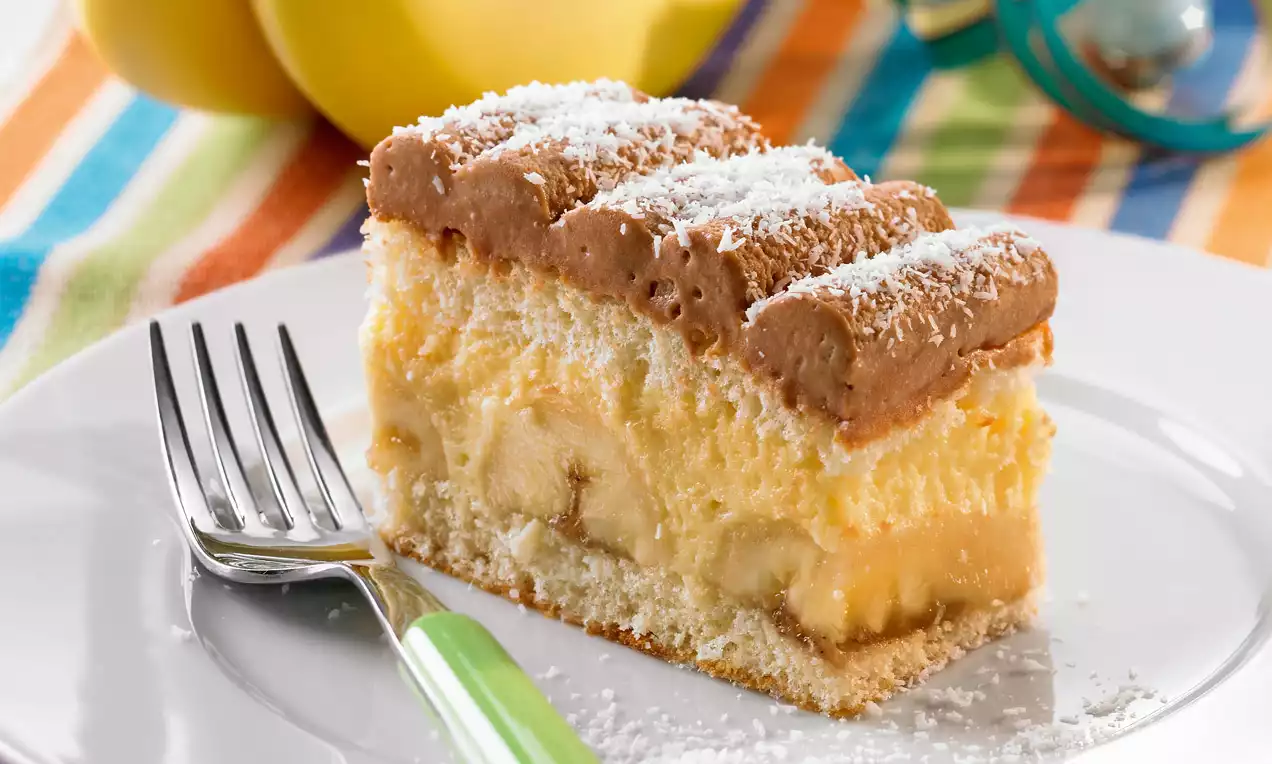 Eine rechteckige Bananen-Puddingschnitte mit hellgelber Füllung, weichem Keksboden und Schokocreme, bestreut mit feinem Kokosraspel auf weißem Teller