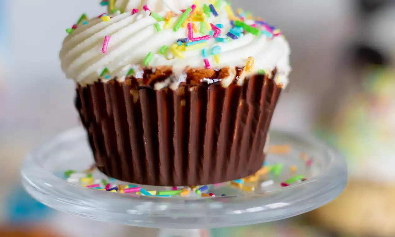 Sladoledni cupcake u tamnosmeđoj čokoladnoj čašici s pahuljastim bijelim vrhom posut šarenim mrvicama na staklenom postolju