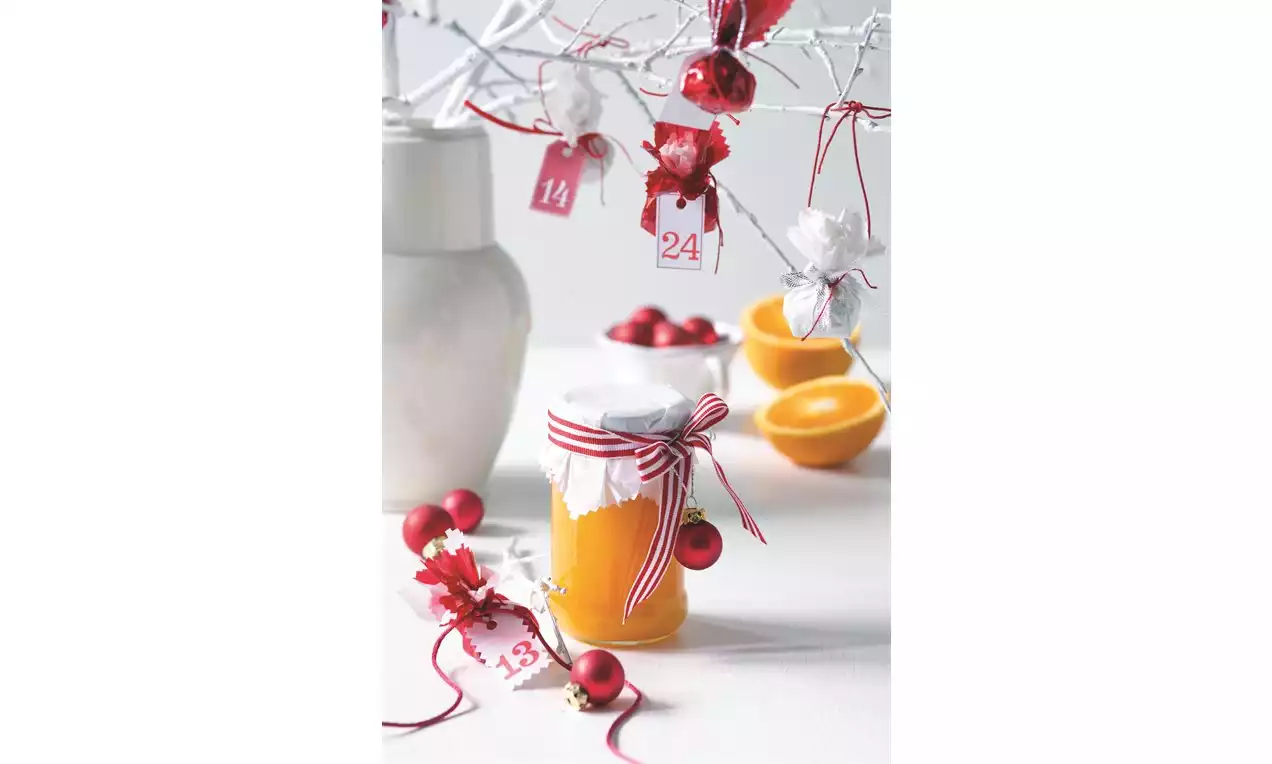 Sklenený pohár naplnený jasne oranžovou marmeládou, uzavretý svetlým papierom a ozdobený červenobielou mašľou, na bielom stole s vianočnými dekoráciami a rozkrojeným pomarančom v pozadí