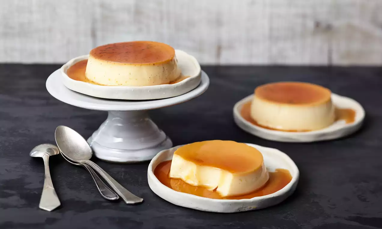 Crème caramel fin și lucios cu strat de caramel auriu, servit pe farfurii albe, alături de tacâmuri metalice pe fundal închis