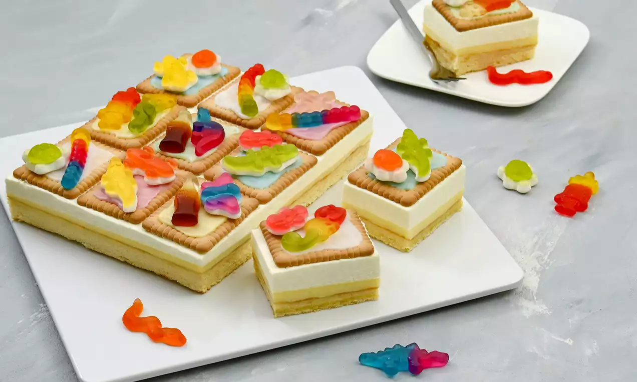 Ein rechteckiger Kuchen mit heller Creme und Keksboden, garnierte Kekse mit bunten Fruchtgummis, auf einem weißen Teller angerichtet