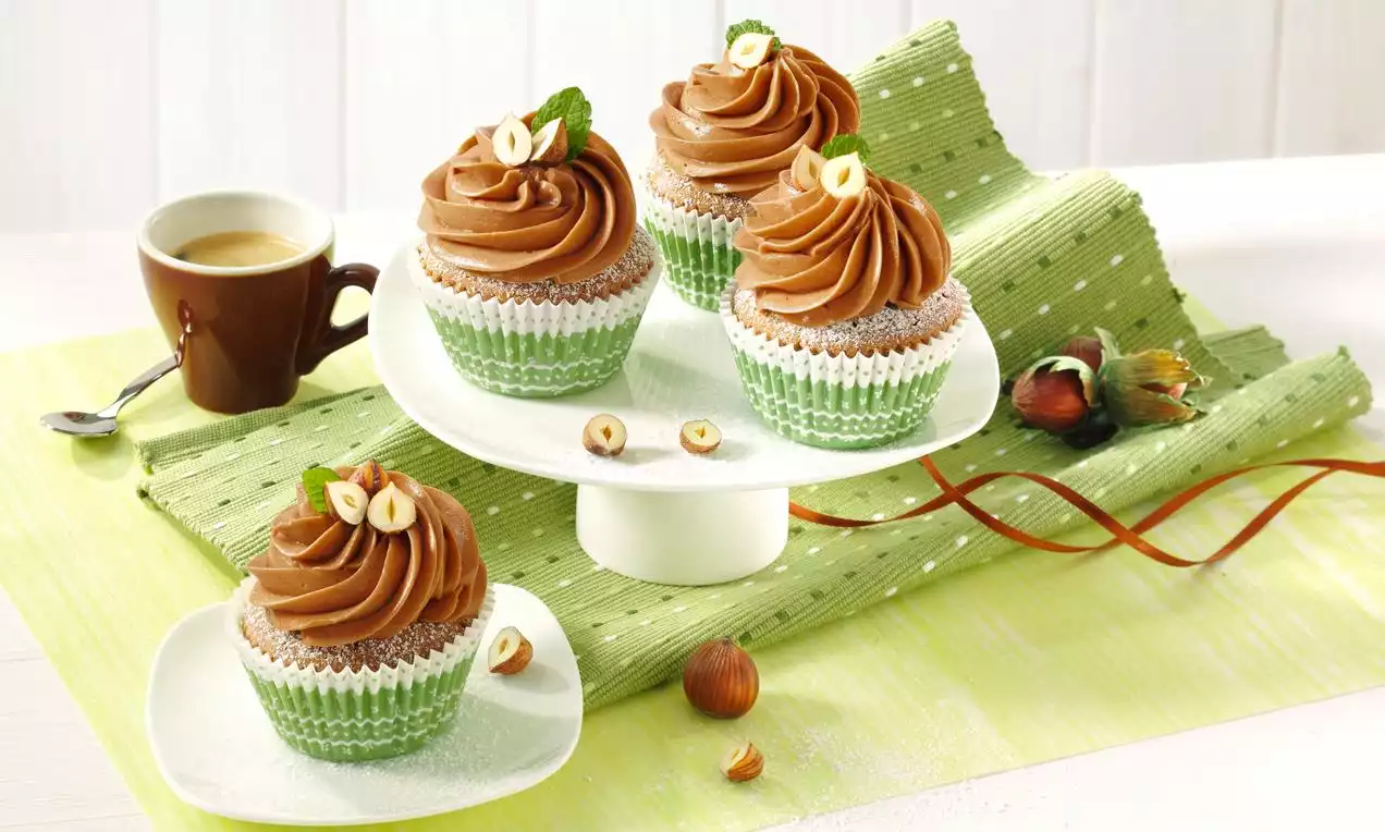 Nugátové muffiny v zelených košíčkoch s vrstvou dohneda krémovej polevy, ozdobené lieskovcami a lístkami mäty na bielom stojane