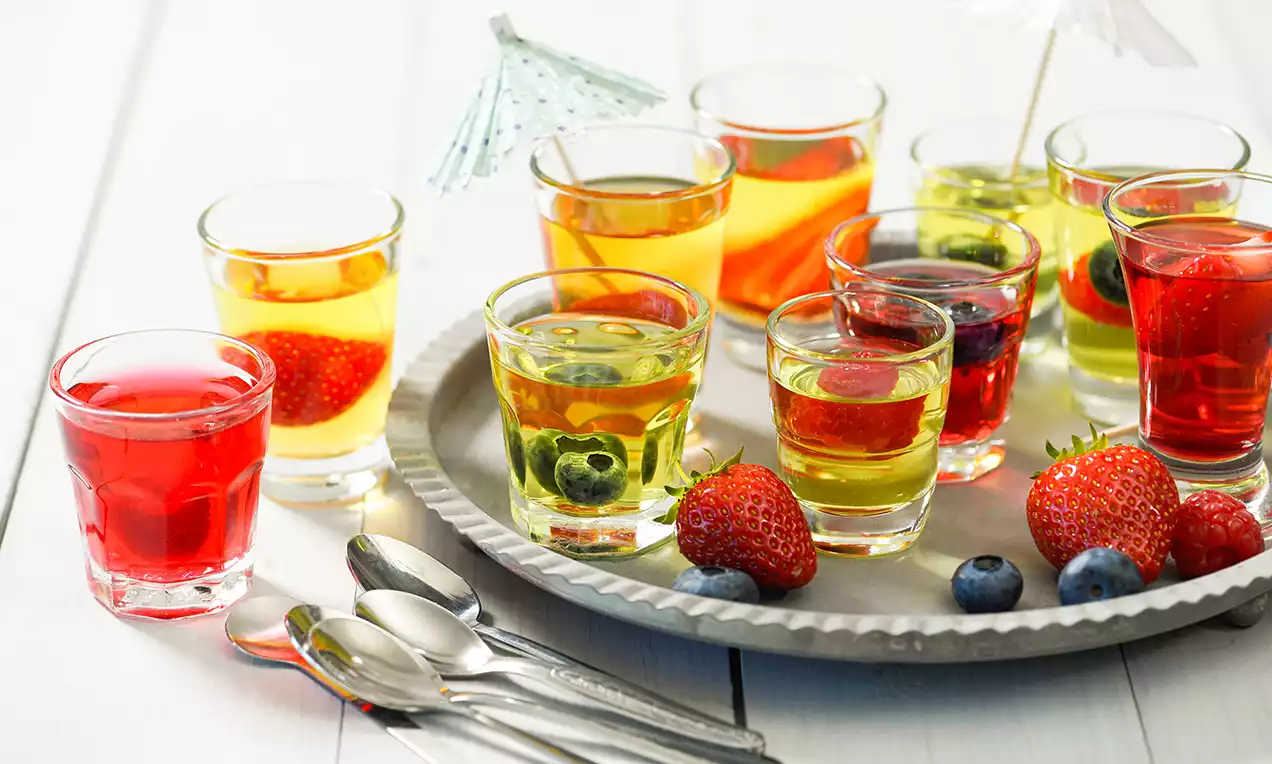Bunte Jelly Shots in klaren Gläsern, mit frischen Beeren und dekorativen Schirmchen auf einem silbernen Teller mit Löffeln drapiert