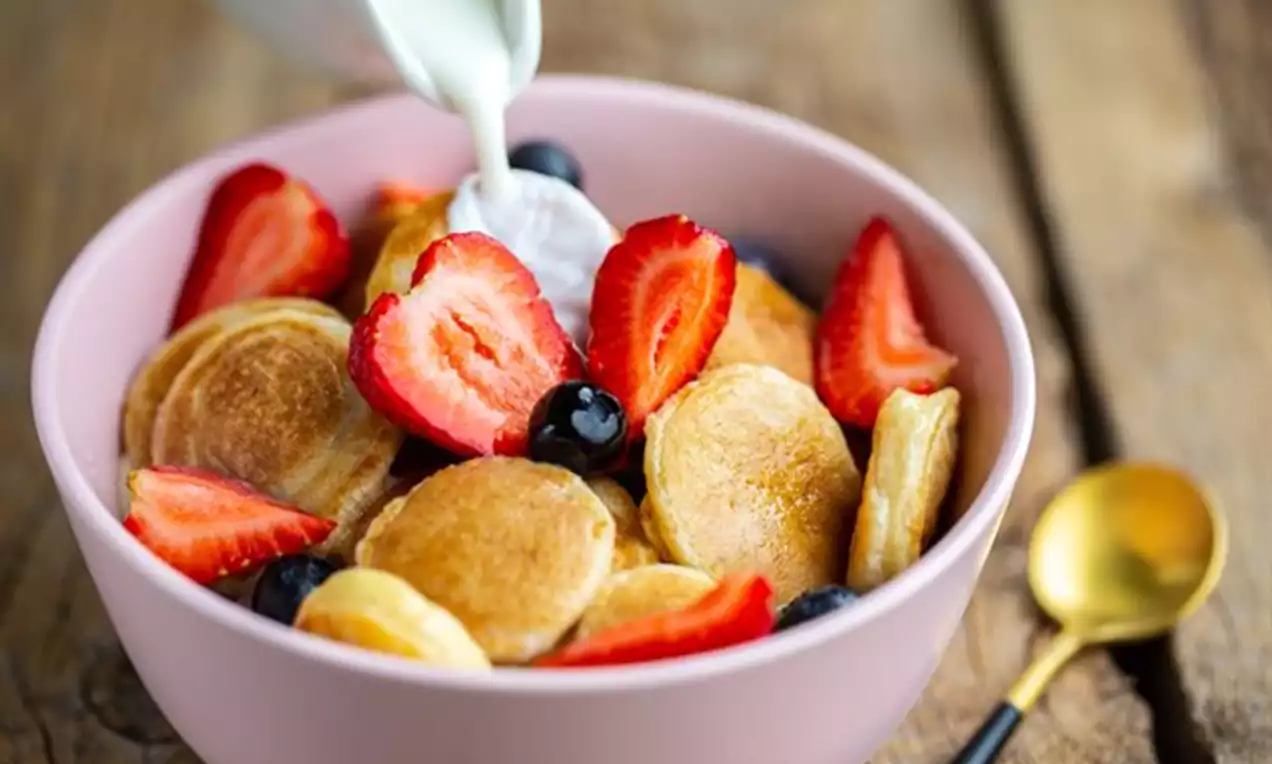 Golden mini pancakes with a soft texture arranged in a pink bowl, topped with fresh sliced strawberries, whole blueberries and pouring cream