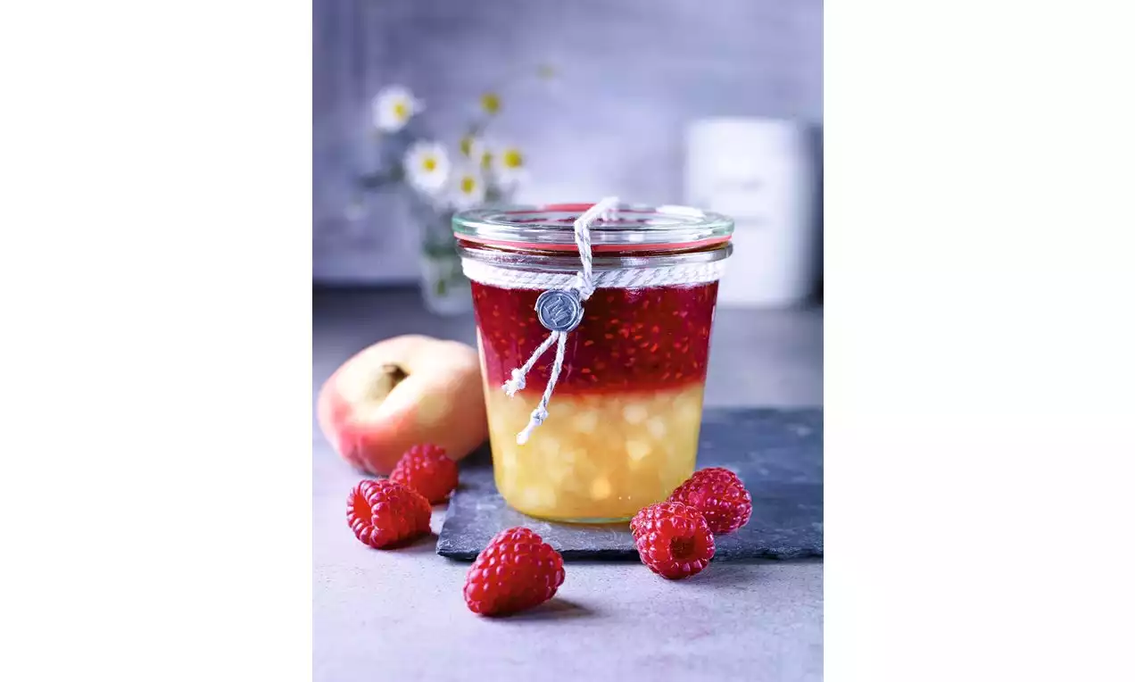 Sklenice s vrstvami broskvového džemu zlatavé barvy a malinového džemu sytě červeného, zdobená provázkem, na tmavém podkladu s malinami