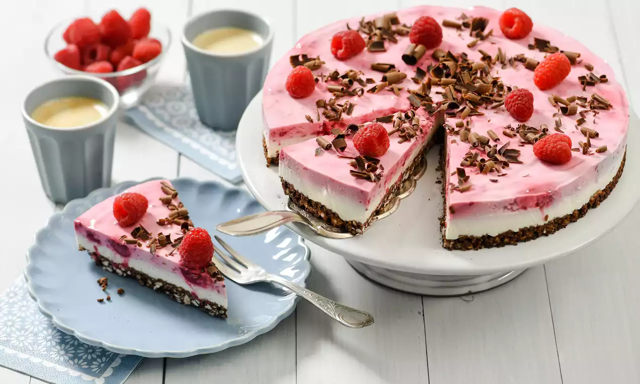 Himbeer-Knusperreis-Torte mit rosafarbenem Himbeerbelag, weißer Cremefüllung, dunklem Knusperboden, garniert mit Himbeeren und Schokospänen auf einer weißen Tortenplatte