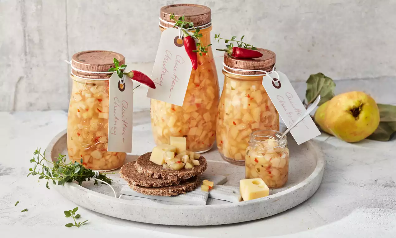 Quitten-Chutney in gläsernen Schraubdeckelbehältern mit roten Chili und frischem Thymian dekoriert auf grauem Tablett mit Brot und Käse