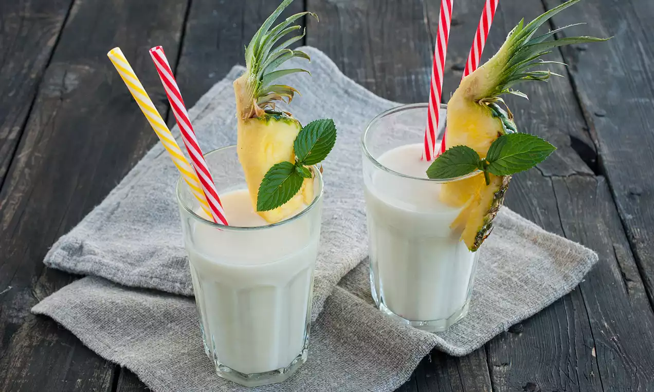 Dva vysoké poháre s krémovo bielym nápojom ozdobené plátkom ananásu, čerstvými lístkami mäty a farebnými slamkami na textílii