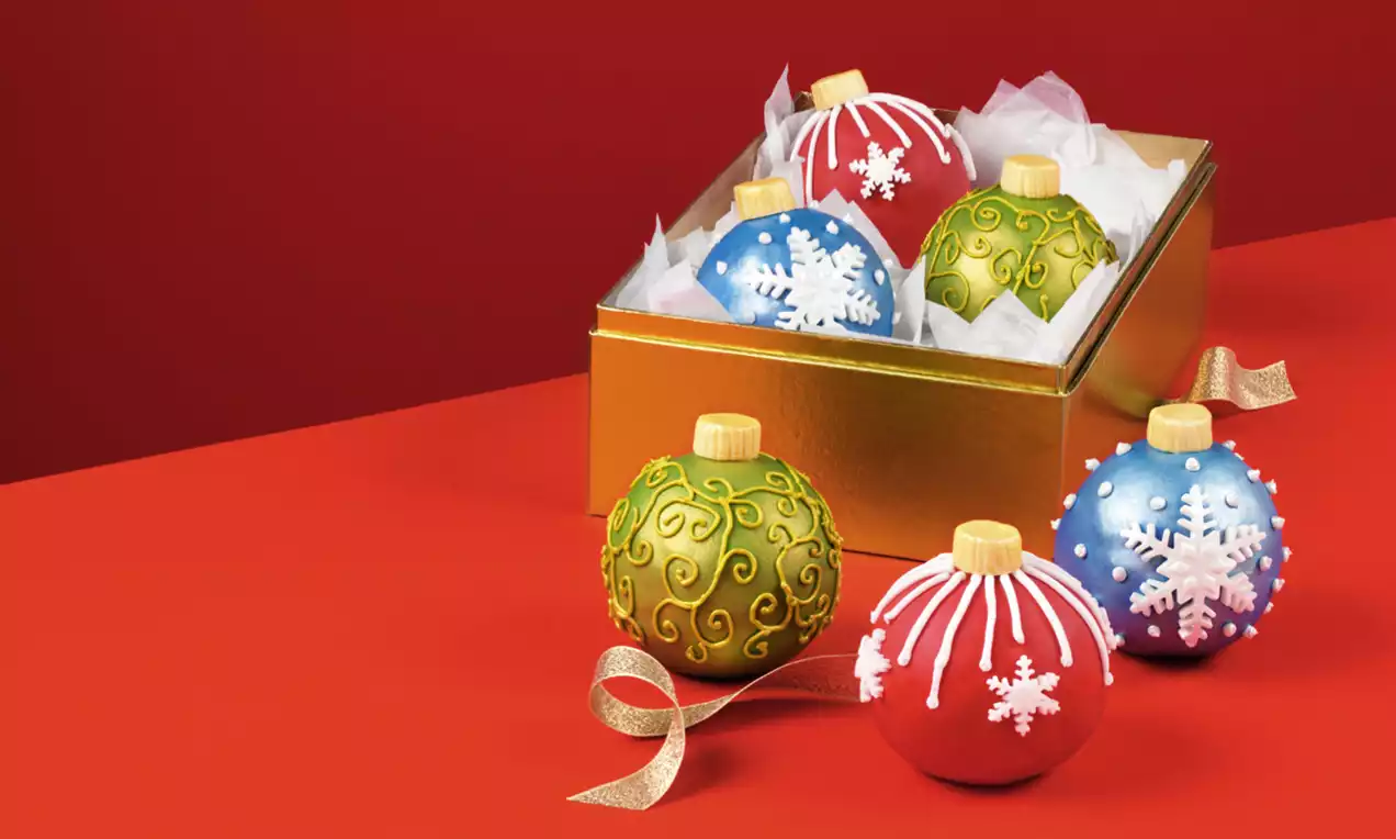 Round bauble-shaped cakes with colorful glossy surfaces, decorative snowflakes, and swirling patterns in a gold box on a red background