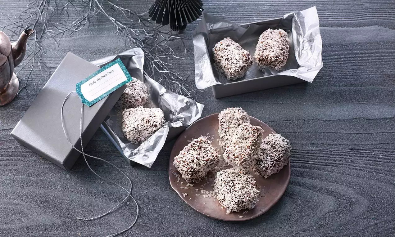 Kleine, würfelförmige Schokoladenkuchen in Kokosflocken gewälzt, teils auf braunem Keramikteller, teils in silberner Geschenkverpackung