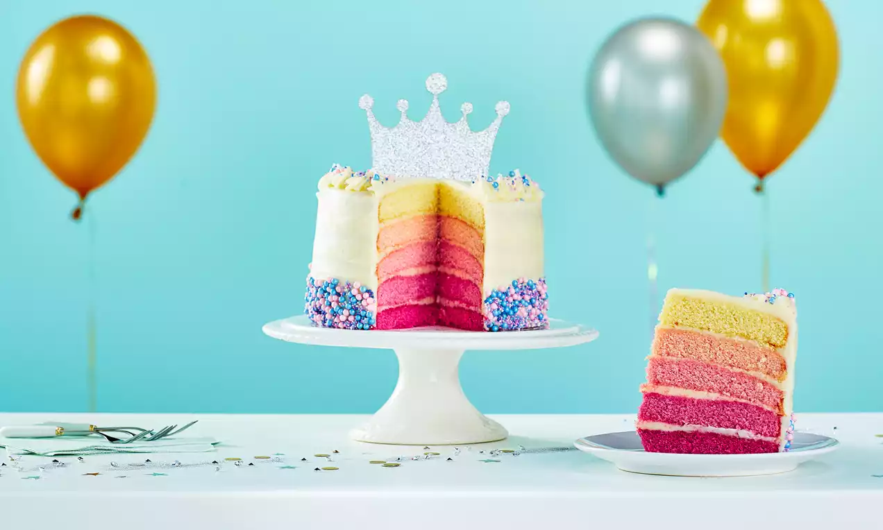 Layered sponge cake with pink, yellow, and coral tones, coated in white frosting with pastel sprinkles, topped with a glittery crown