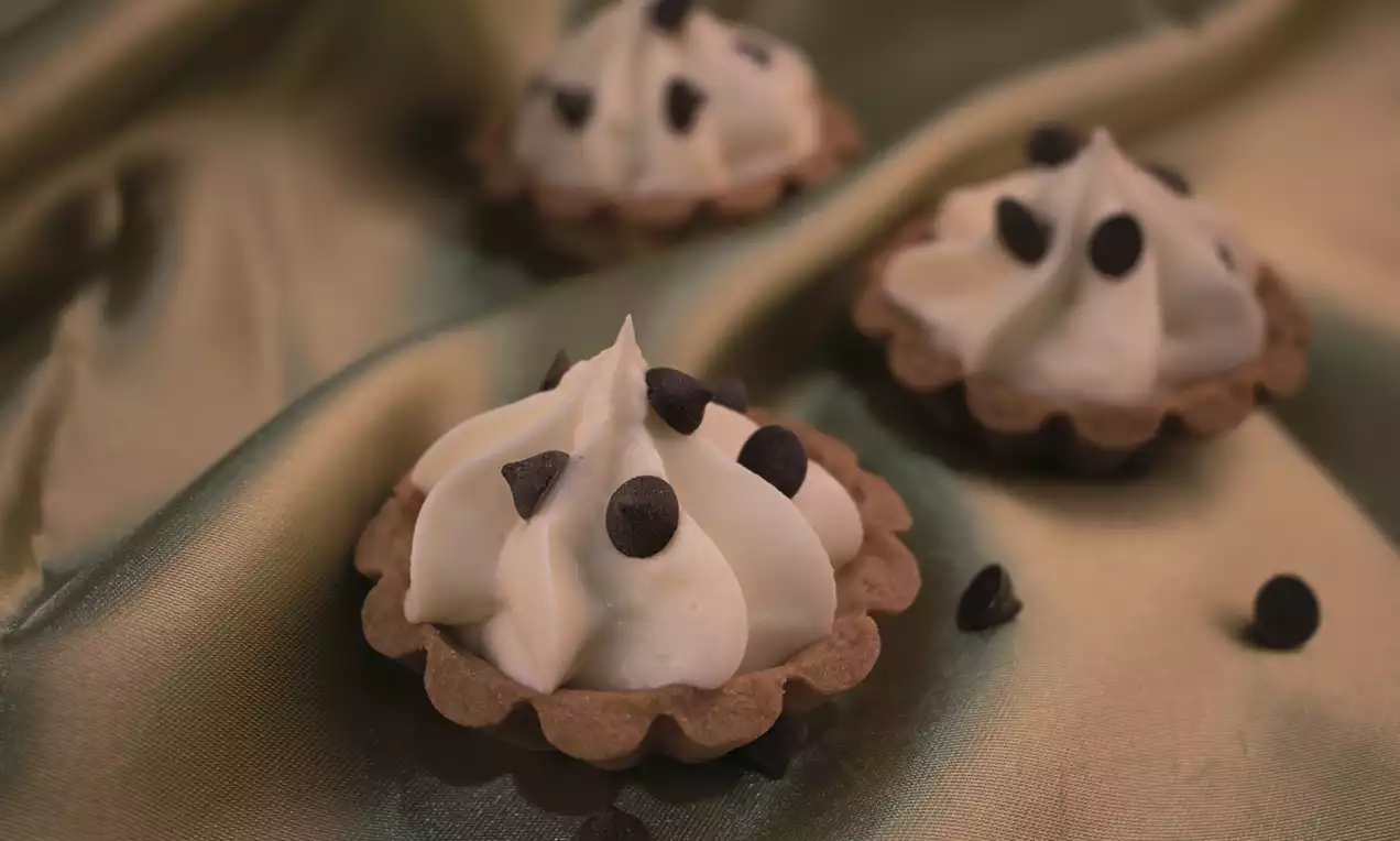 Golden brown tart shells filled with smooth swirls of pale cream, topped with scattered dark chocolate chips on a silky fabric background