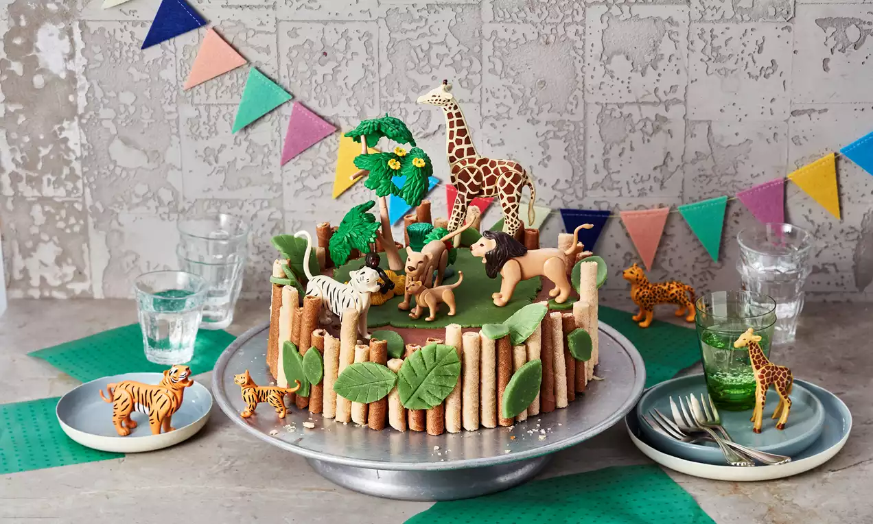 Runde Torte mit grünem Fondant, gesäumt von Keksstäben, dekoriert mit Blättern, Kekstieren und Baum-Elementen auf einer silbernen Platte