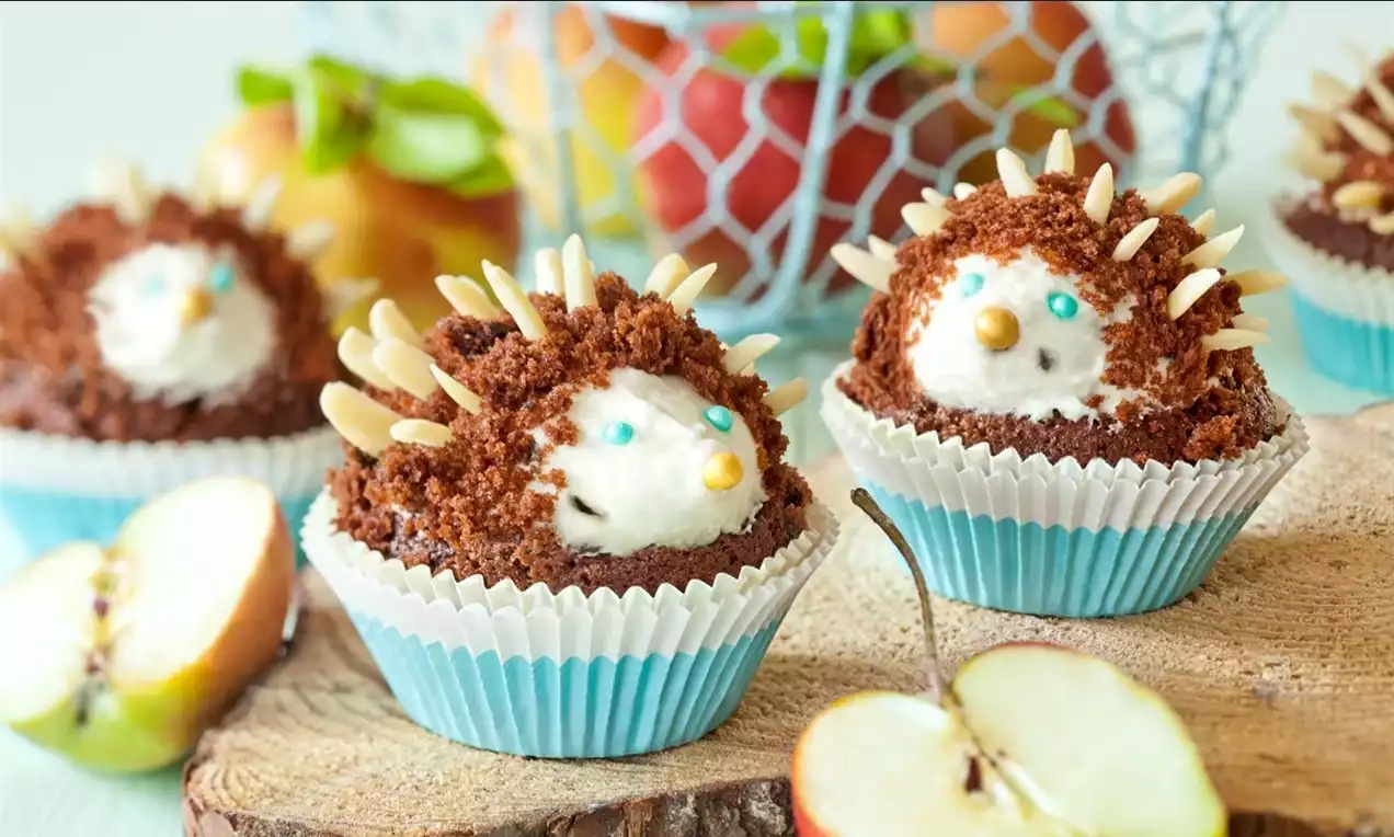 Kakaový muffin s krtkovým krémom ozdobený mandľovými pichliačmi, svetlomodrý košíček vedľa jablka na drevenom podnose.