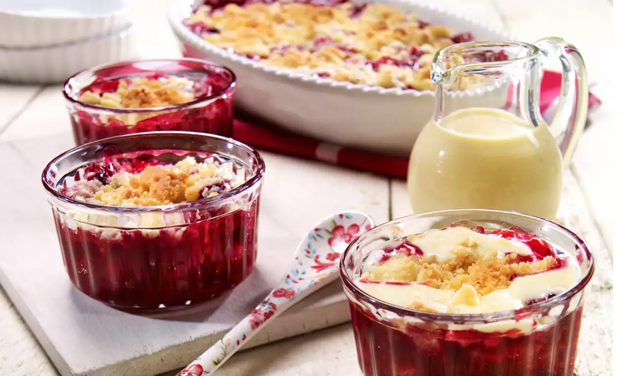 Rote Beeren mit glänzender Oberfläche unter goldbraunen Crumble-Streuseln in Glasförmchen, daneben eine Glas-Karaffe mit heller Vanillesoße