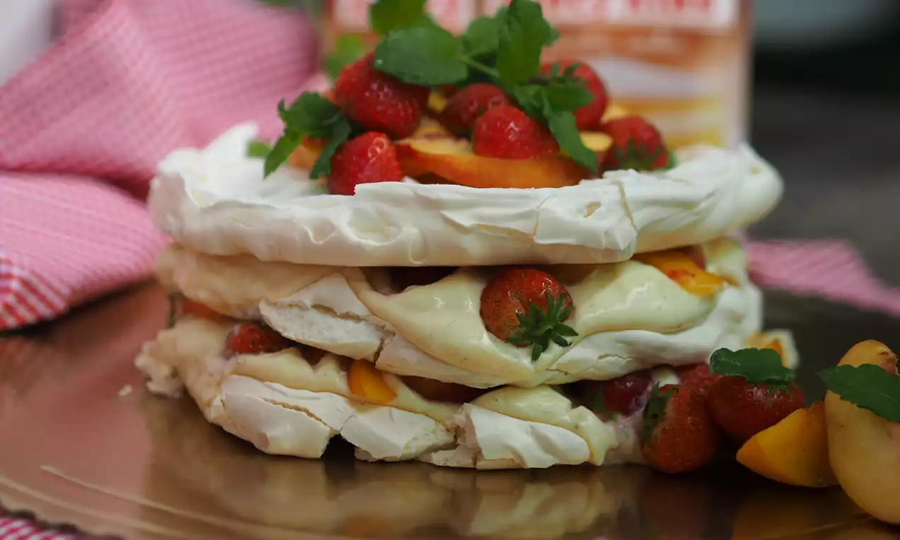 Pavlova dort s bílými křupavými vrstvami, krémem mezi nimi, zdobený čerstvými jahodami, lístky máty a plátky broskví na zlatém podnose