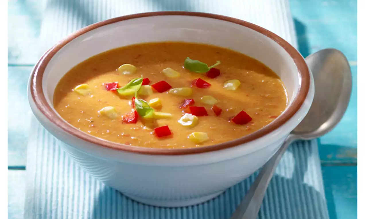 Paprika-Mais-Suppe cremig-rot mit gelben Maiskörnern, grünen Kräuterflocken und einem Stück Bauernbrot in weißer Schale