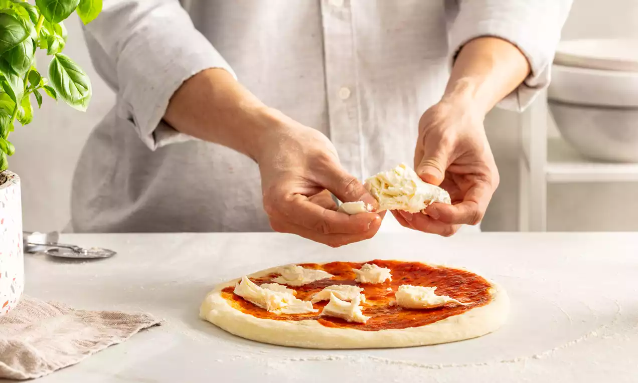 Hands placing small pieces of soft white cheese onto a round sourdough pizza base spread with vibrant red tomato sauce on a floured surface