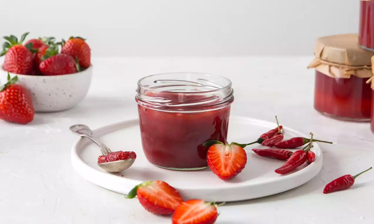 Jahodový džem sýtočervenej farby v sklenenom pohári na bielom tanieri, obklopený čerstvými jahodami, chilli papričkami a lyžičkou s džemom